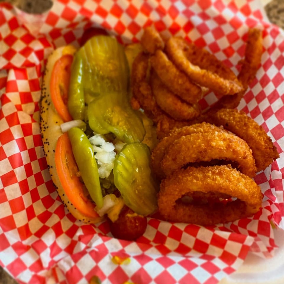 Mugsy Dogs hot dog with tomatoes, pickles, and onions, served with a side of onion rings.