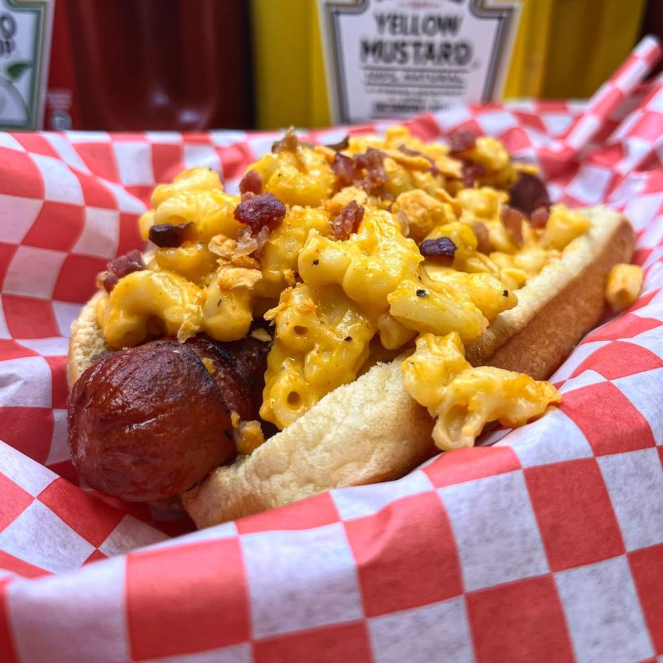 Mugsy Dogs' hot dog with macaroni and cheese, bacon bits, and crispy onions in a red checkered basket.