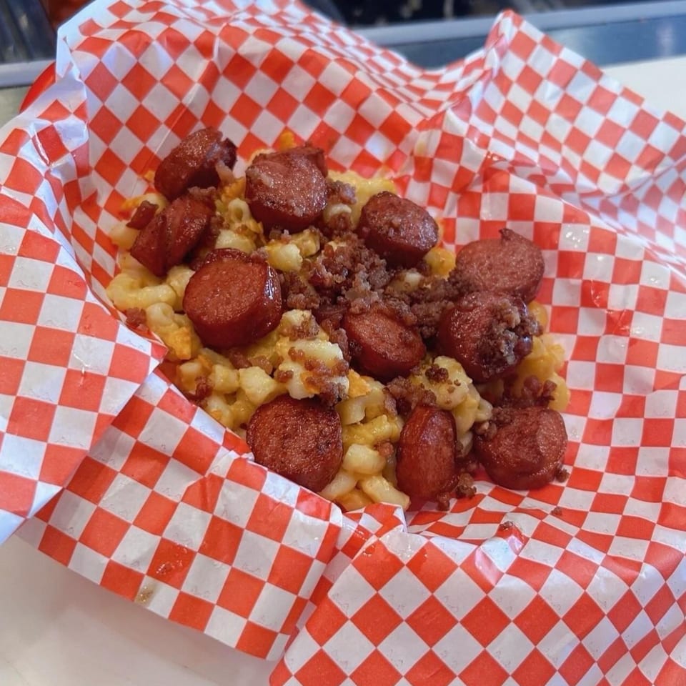 Mugsy Dogs' mac and cheese topped with sliced hot dogs and bacon bits in a red checkered basket.