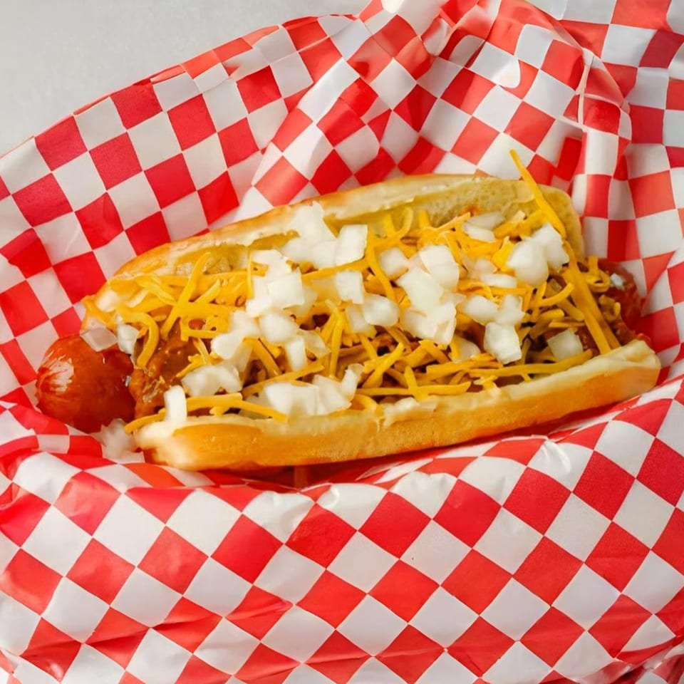 Mugsy Dogs' chili cheese hot dog with diced onions in a red and white checkered paper tray.