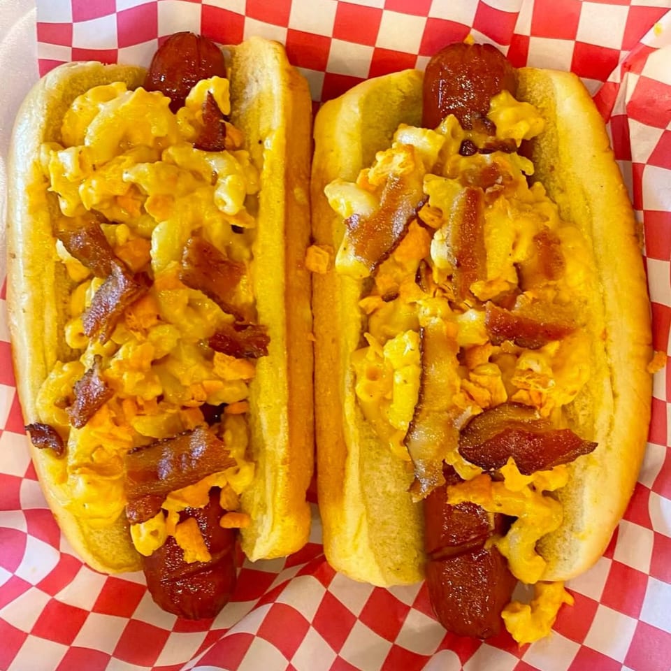 Two Mugsy Dogs hot dogs with macaroni and cheese, bacon, and cheese puffs in red checkered paper.