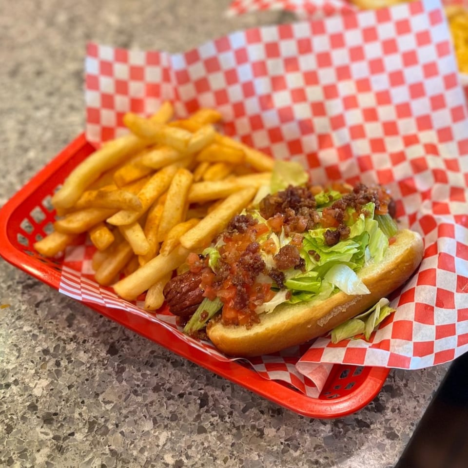 Mugsy Dogs hot dog with lettuce, bacon, and tomatoes, served with fries in a red basket.
