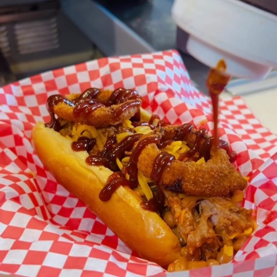 Mugsy Dogs' hot dog with pulled pork, cheese, onion rings, and BBQ sauce in a red checkered basket.