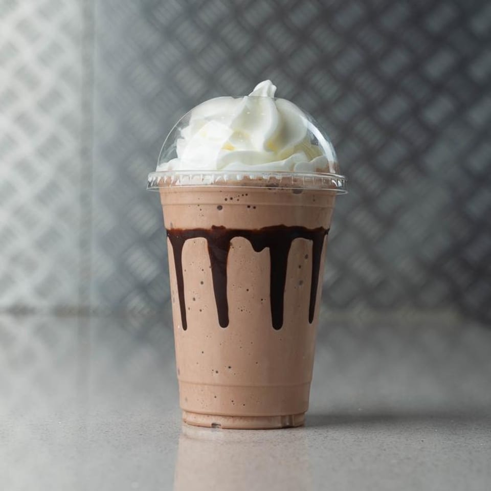 Mugsy Dogs chocolate milkshake with whipped cream and chocolate drizzle in a clear plastic cup.