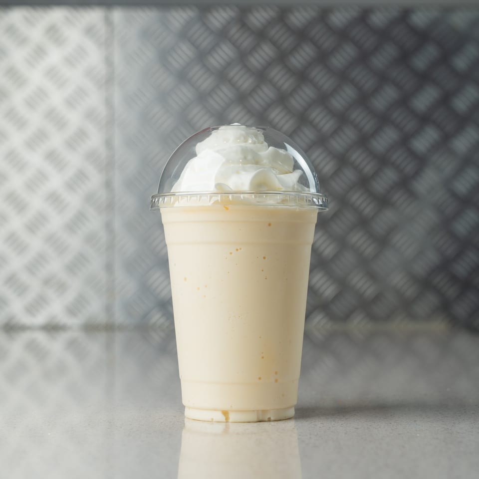 A creamy vanilla milkshake with whipped cream in a clear plastic cup against a metal background.