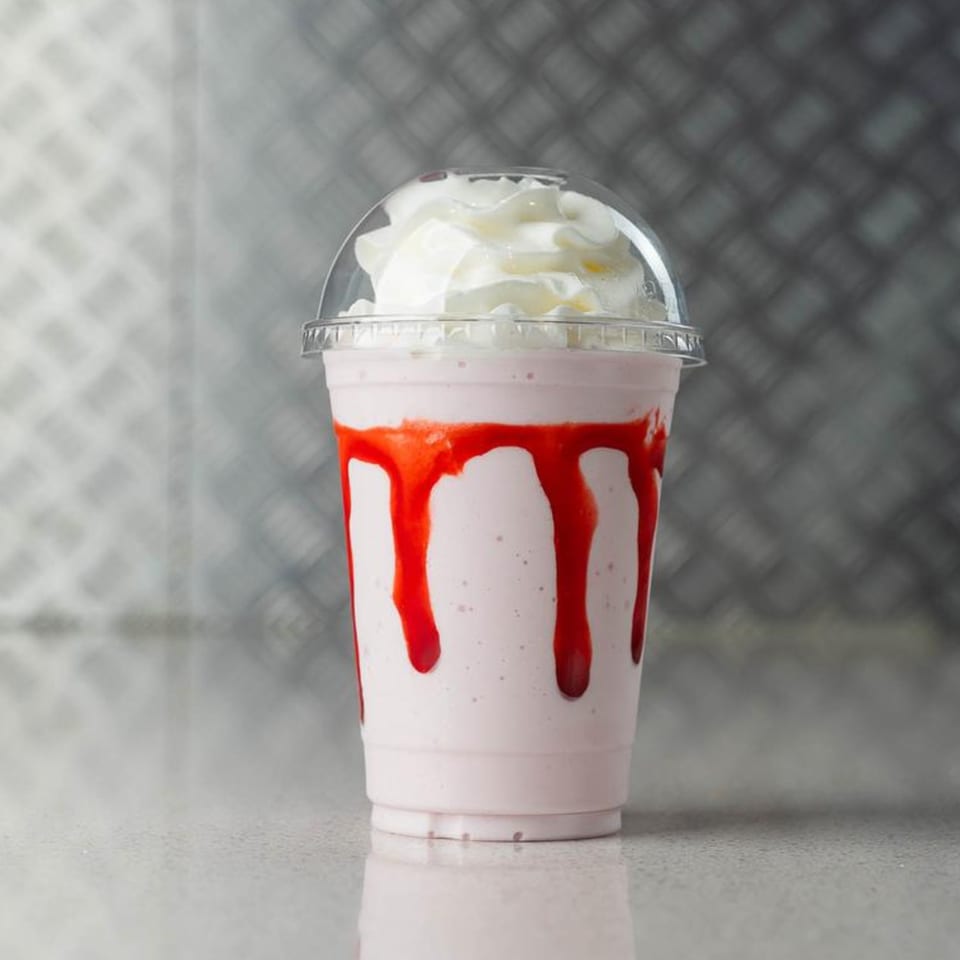 Strawberry milkshake with whipped cream and red syrup drizzle in a clear plastic cup.